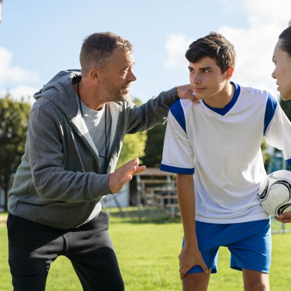 Mature coach teaching strategy to high school team on football field. Young soccer players standing together united and listening coach motivational speech. Coach giving team advise before school match.